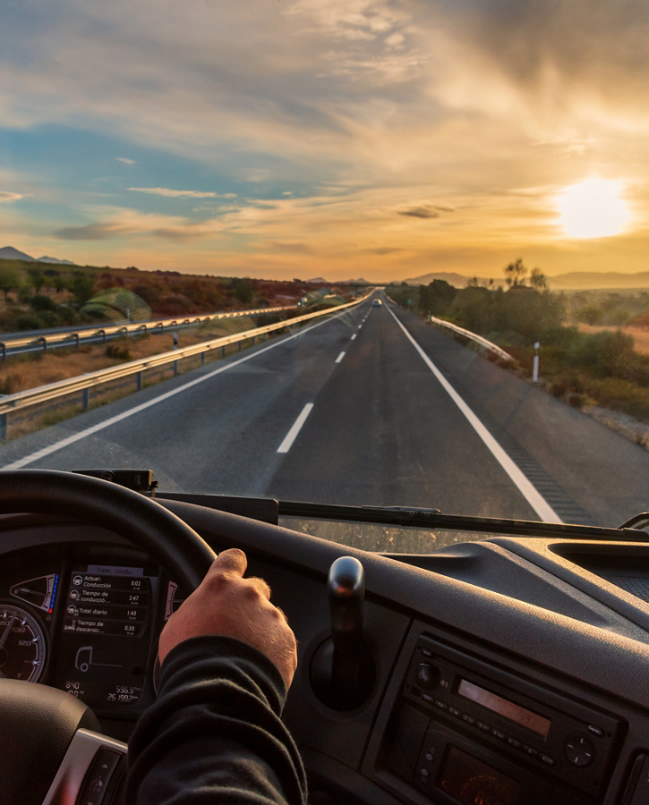  View from inside a truck cabin as it drives down a highway at sunset, with the driver's hands on the steering wheel. The sun is setting over the horizon, casting a warm glow on the open road, which is flanked by guardrails and sparse vegetation.