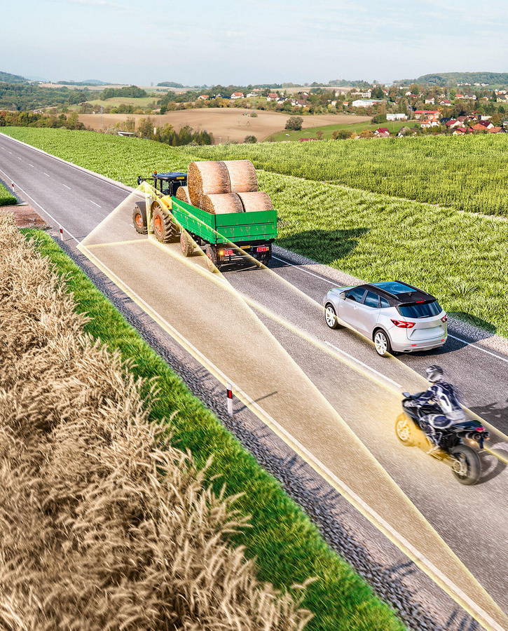 Schematische Darstellung eines Traktors mit einem Anhänger, der auf einer Landstraße fährt. Der Anhänger ist mit zylindrischen Strohballen beladen. Hinter dem Gespann fährt ein Pkw und dahinter, leicht nach links versetzt auf der Gegenfahrbahn, ein Motorrad. Der Traktor scannt mit gelben Radarstrahlen die Gegenfahrbahn ab und erkennt in einigem Abstand das Motorrad.