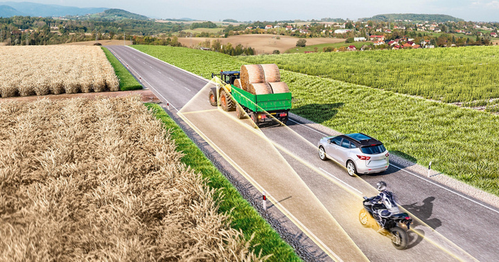 Schematische Darstellung eines Traktors mit einem Anhänger, der auf einer Landstraße fährt. Der Anhänger ist mit zylindrischen Strohballen beladen. Hinter dem Gespann fährt ein Pkw und dahinter, leicht nach links versetzt auf der Gegenfahrbahn, ein Motorrad. Der Traktor scannt mit gelben Radarstrahlen die Gegenfahrbahn ab und erkennt in einigem Abstand das Motorrad.