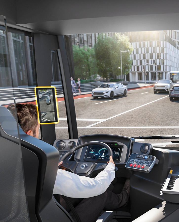 View of a city bus’ interior looking through the windshield onto a city street. The driver is sitting on the left behind the steering wheel. There is a camera monitor system mounted on the A-columns with two monitors on the right side and one on the left side. The monitors show what is happening around the bus from different angles.