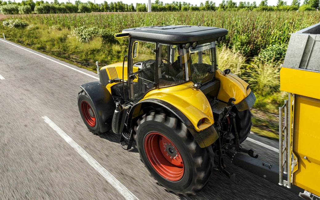 A yellow tractor with red wheels is pulling a trailer on a paved road, surrounded by lush green fields and vegetation on a sunny day. The tractor is viewed from behind, showcasing its robust build and large tires.