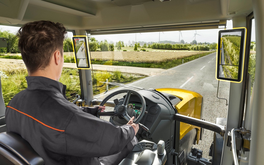 A person with short brown hair, wearing a gray and orange jacket, is driving a yellow tractor on a rural road. The view through the windshield shows fields and wind turbines in the distance under a partly cloudy sky. The interior of the tractor cabin is visible.