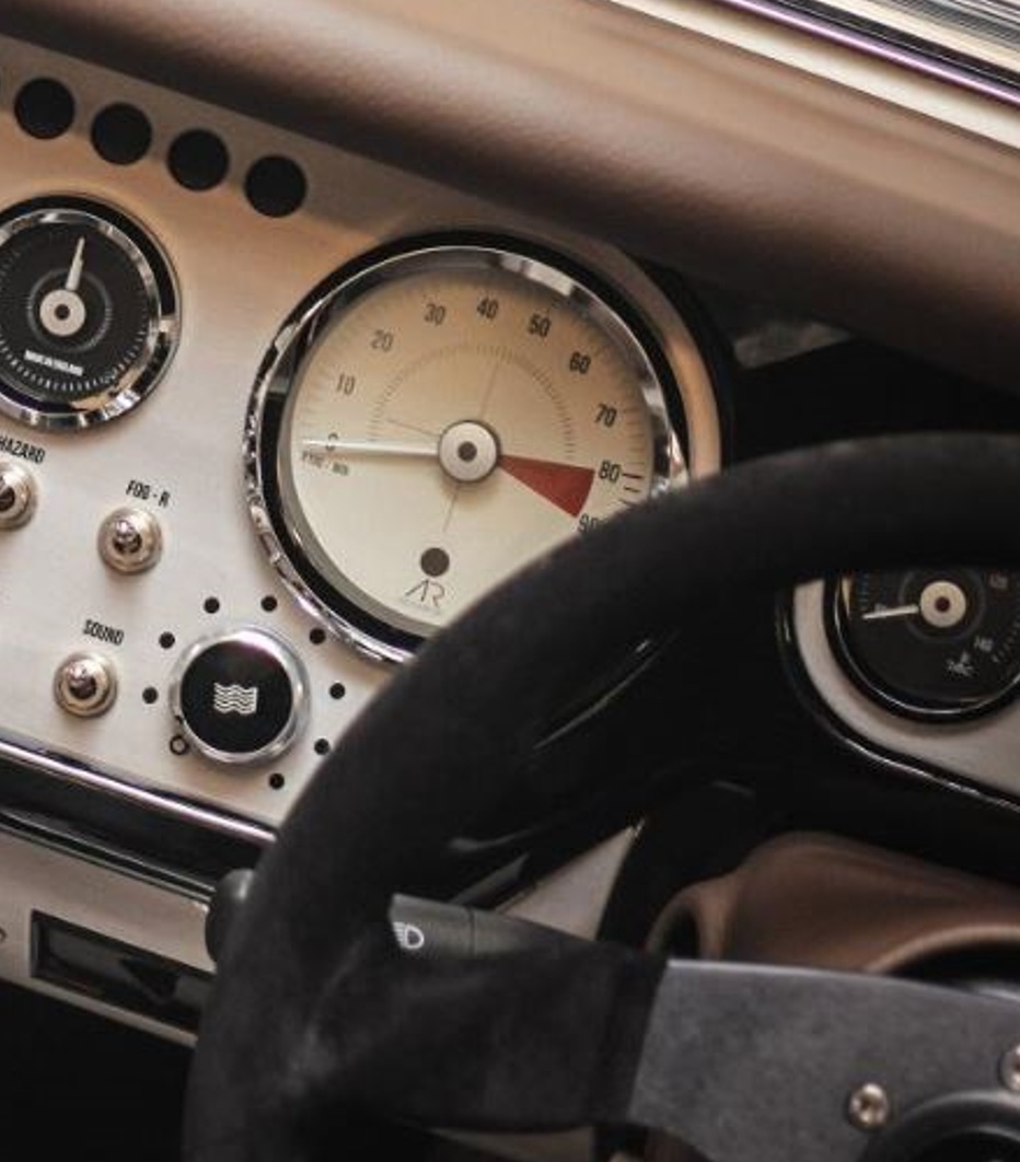  Close-up view of a classic car dashboard, featuring a speedometer with the needle pointing at around 65, a temperature gauge, and other controls. The dashboard has a vintage design with metallic accents and a wooden steering wheel partially visible.