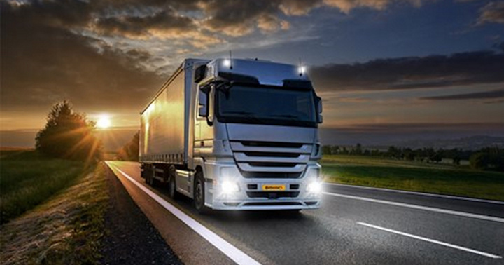 A silver truck with a trailer viewed from the front is driving along a country road at dusk. The headlights shine brightly. The sun is setting in the background.