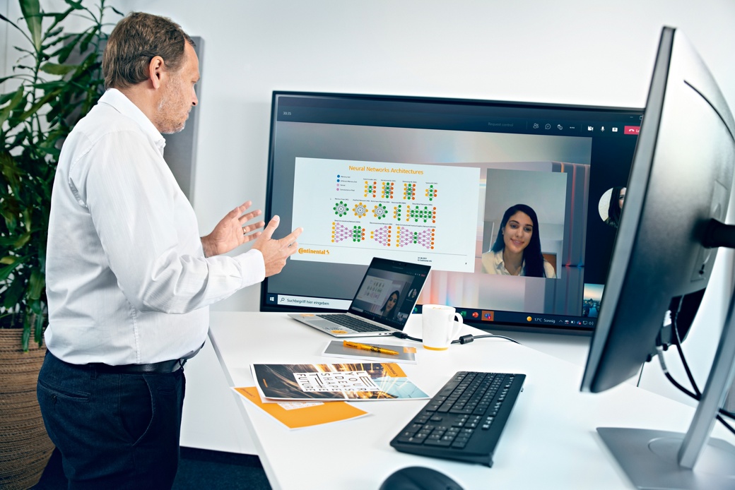 An older man in a white shirt stands at a standing workstation with keyboard, screen and laptop and looks at a TV screen on which a video conference is taking place