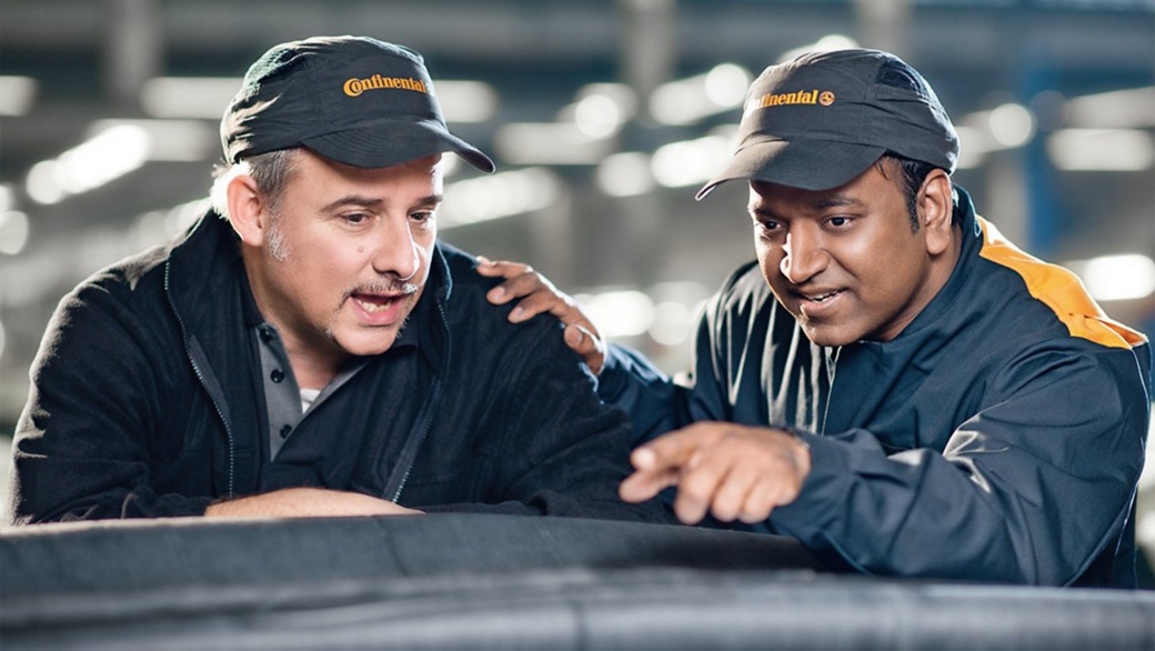An older and a younger man in work clothes and caps with Continental logo look at a conveyor belt