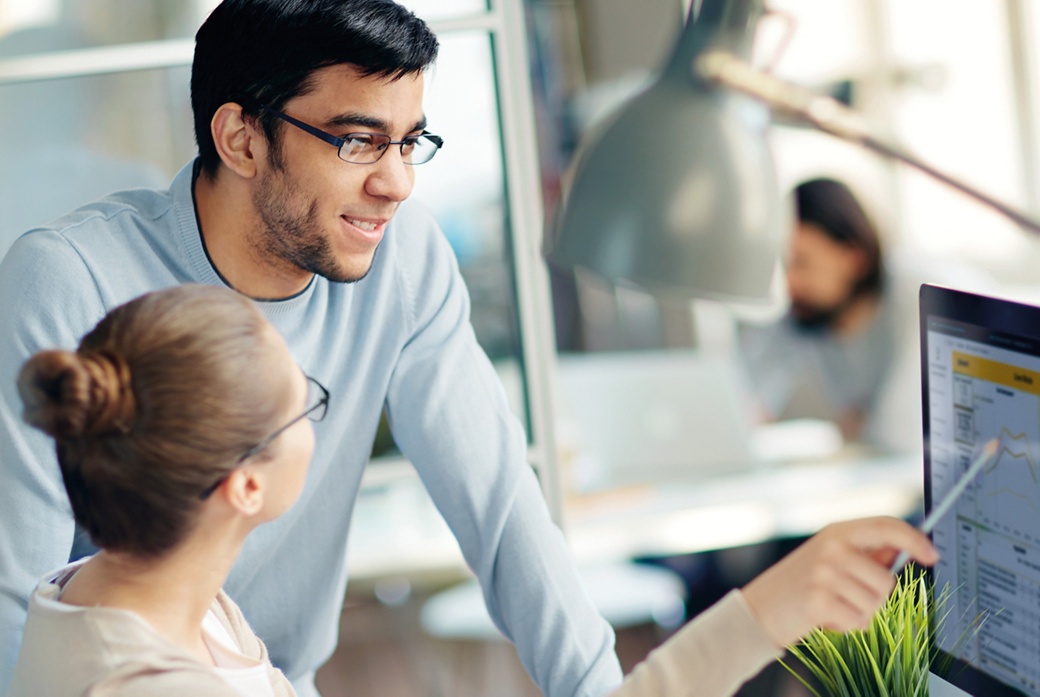A man with glasses is standing at a desk looking at what a woman with glasses is showing him sitting on a display