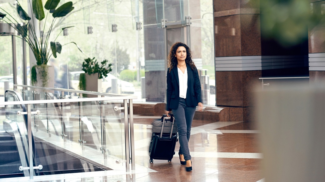 A woman in a jacket, jeans and pumps pulls a trolli behind her as she walks through a reception hall with an escalator