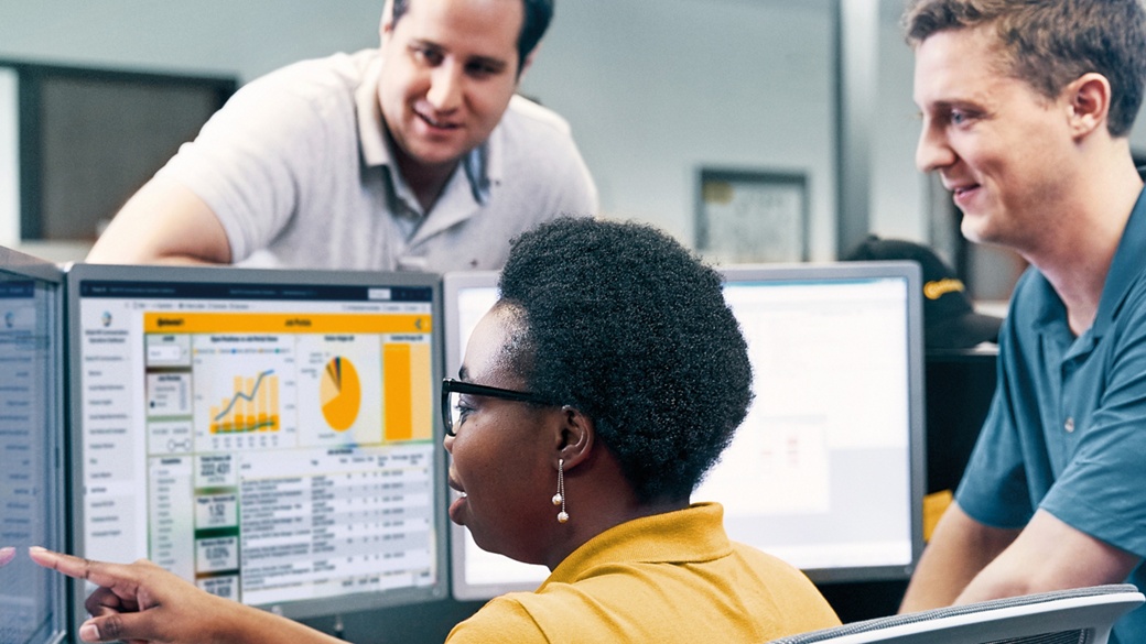 A middle-aged woman sits in front of three PC screens displaying statistics and shows two men something on the left display.