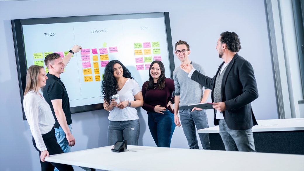 Three young women and two young men stand in front of a white board and look towards a middle-aged man in a jacket who explains the contents of the white board